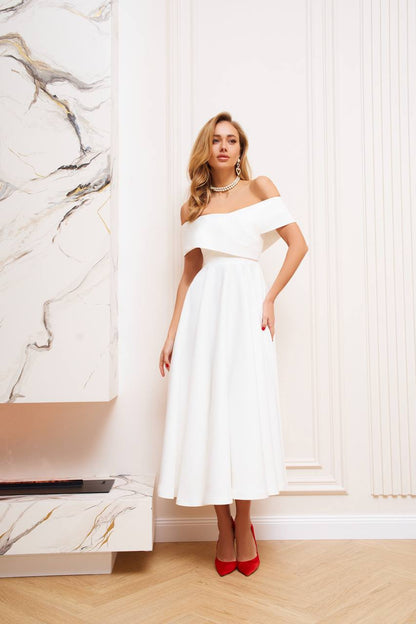 Woman in a white off-shoulder dress standing in a stylish interior setting.