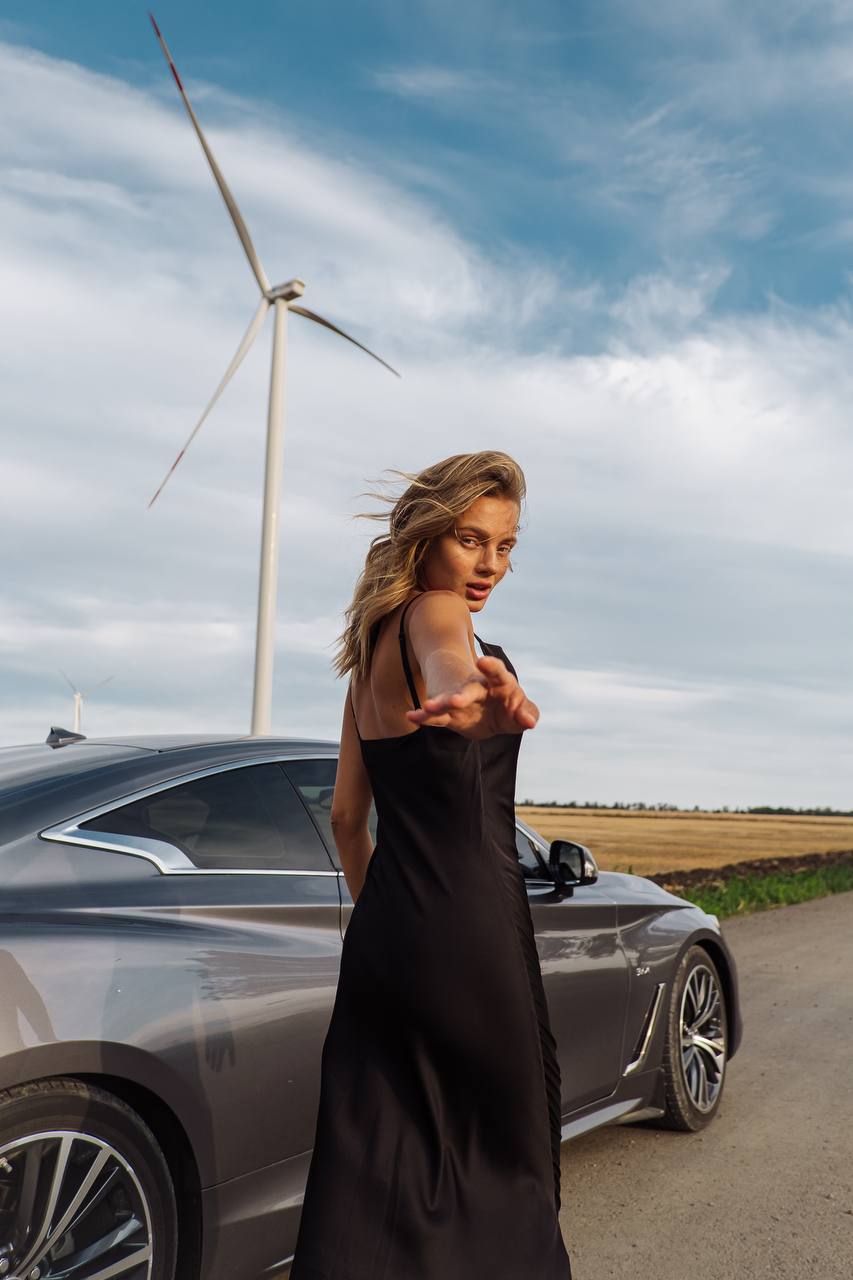 Woman in a black dress standing next to a car with a wind turbine in the background