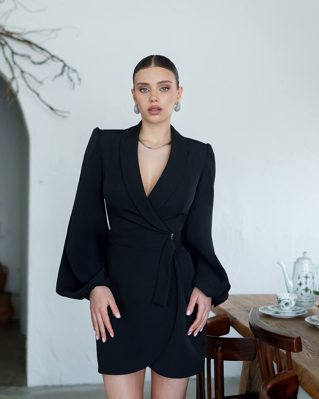Woman wearing a black dress standing in a room with a white wall and wooden furniture.