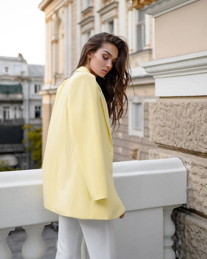 A woman wearing a light yellow oversized blazer and white pants, standing on a balcony with a classical building in the background.