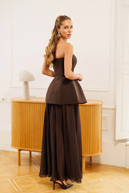 A woman modeling a dark brown strapless gown with a structured peplum waist and a flowing chiffon skirt, standing in a bright room with a wooden cabinet and light-colored walls.