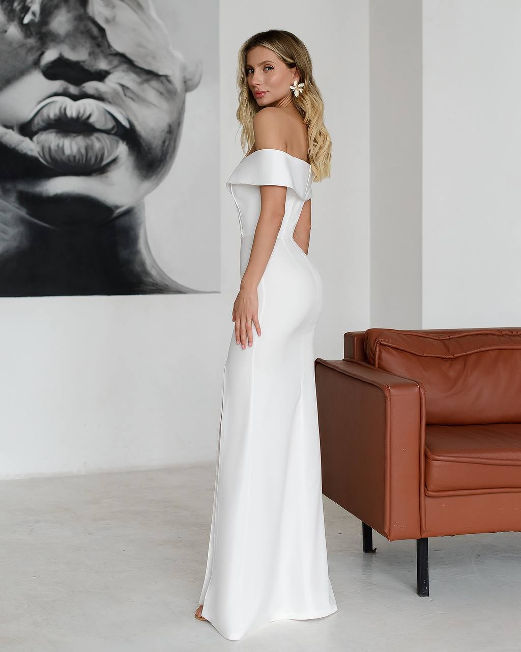 Woman in a white off-shoulder gown standing in a room with a brown leather couch and a large black and white portrait.