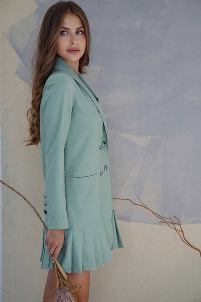 Woman wearing a light green blazer and pleated skirt against a neutral background