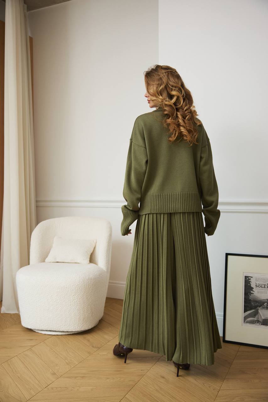 Woman in a green outfit standing in a room with a white chair and framed picture.