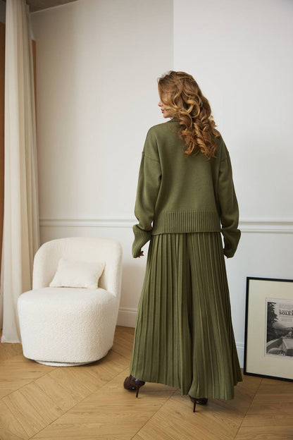 Woman in a green outfit standing in a room with a white chair and framed picture.
