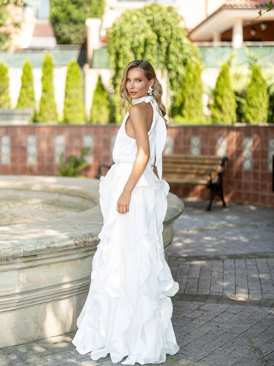 A woman wearing a white sleeveless wedding gown with a ruffled tiered skirt, standing outdoors in a garden courtyard setting.