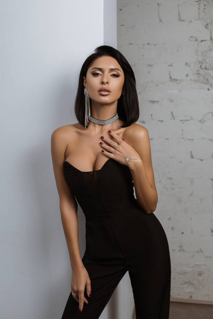 Woman in a black one-shoulder jumpsuit posing against a textured wall.