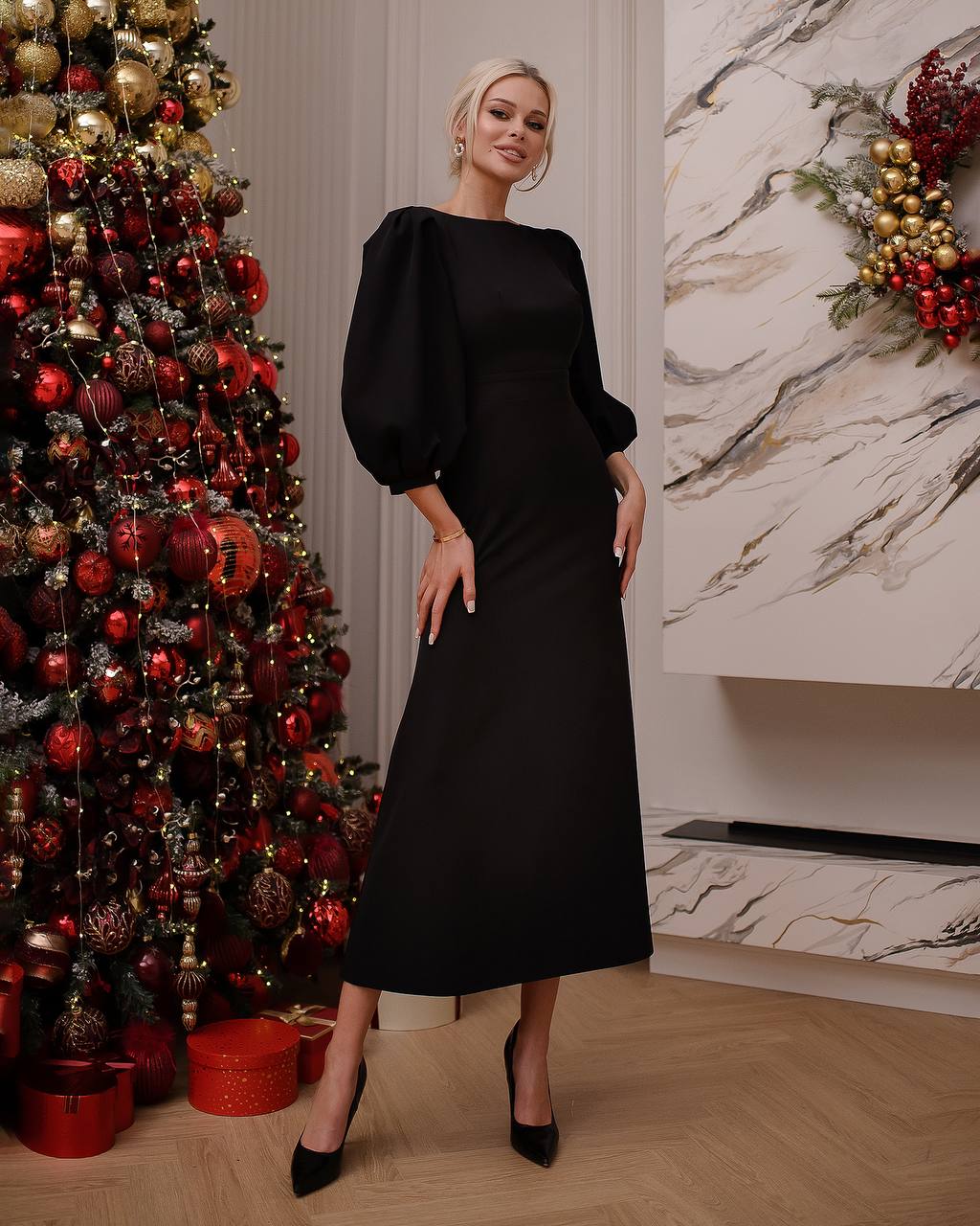 Woman in a black dress standing next to a decorated Christmas tree indoors.