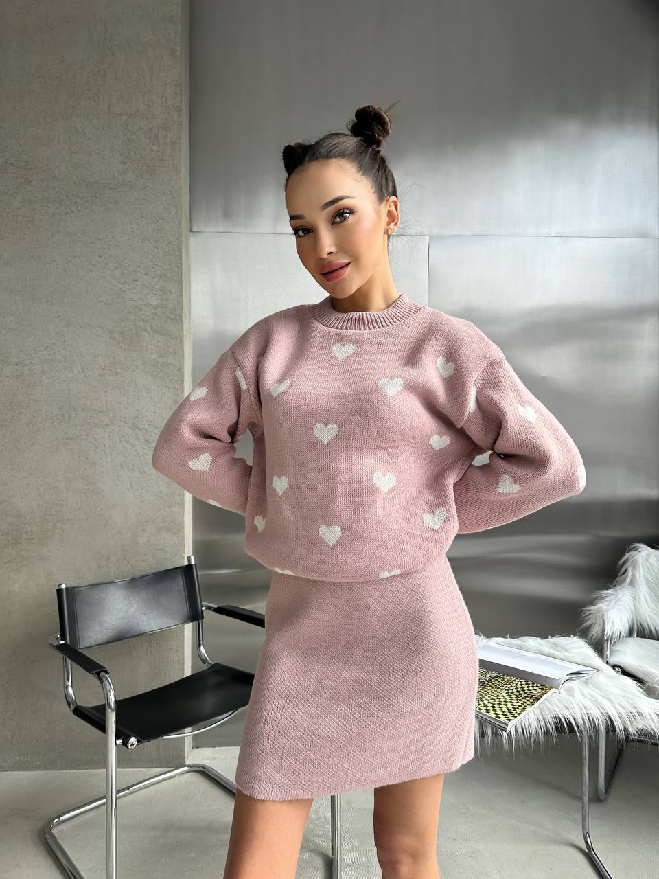 Woman wearing a pink sweater with heart patterns and matching skirt in a minimalistic room.