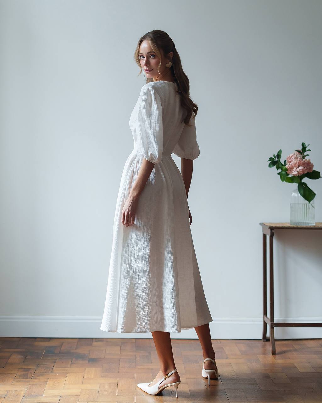 Woman in a white dress standing in a room with a wooden floor and a vase of flowers on a table.