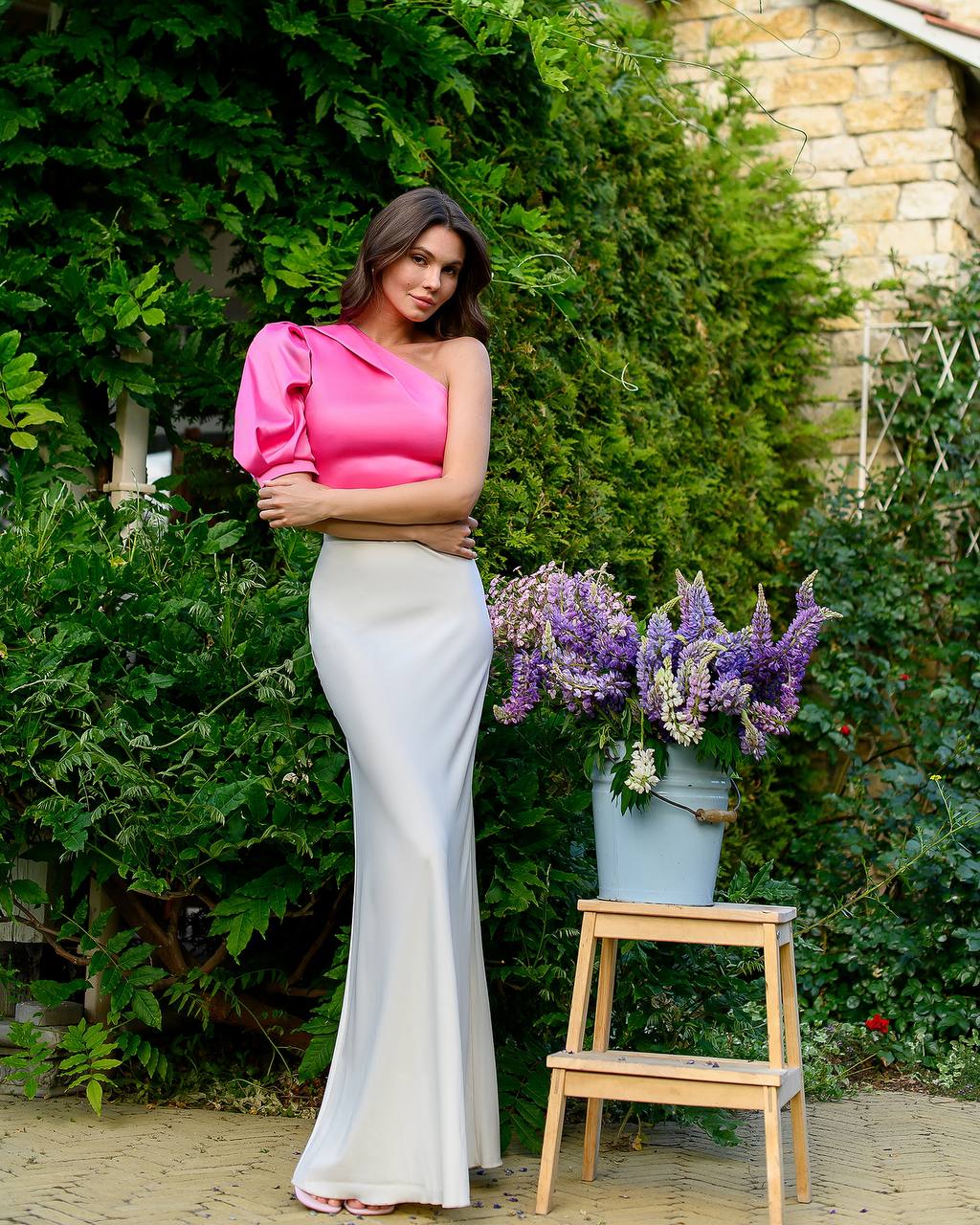 Woman in a pink top and white skirt standing next to a wooden stool with flowers outdoors.