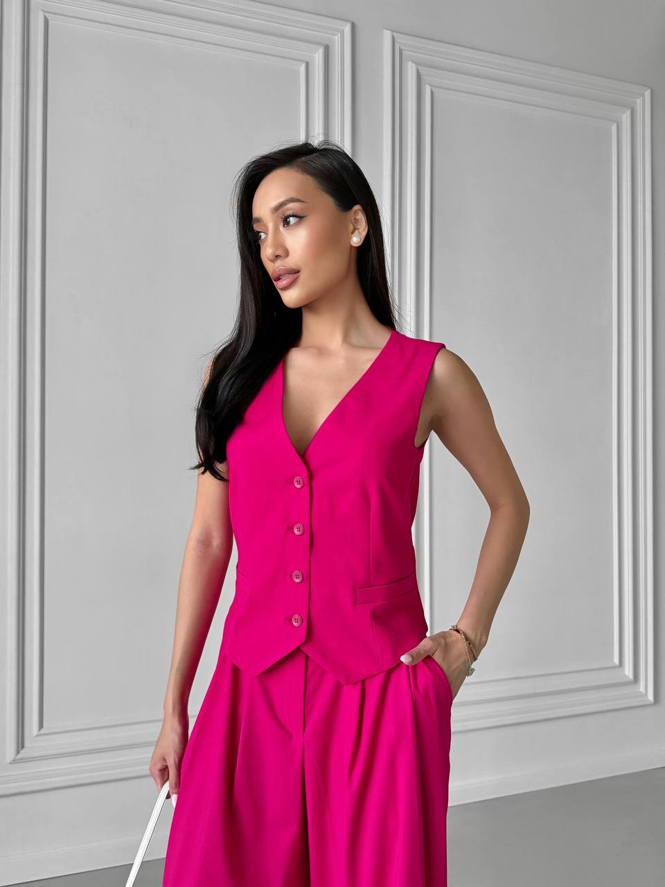 Woman wearing a bright pink sleeveless jumpsuit against a gray paneled wall.