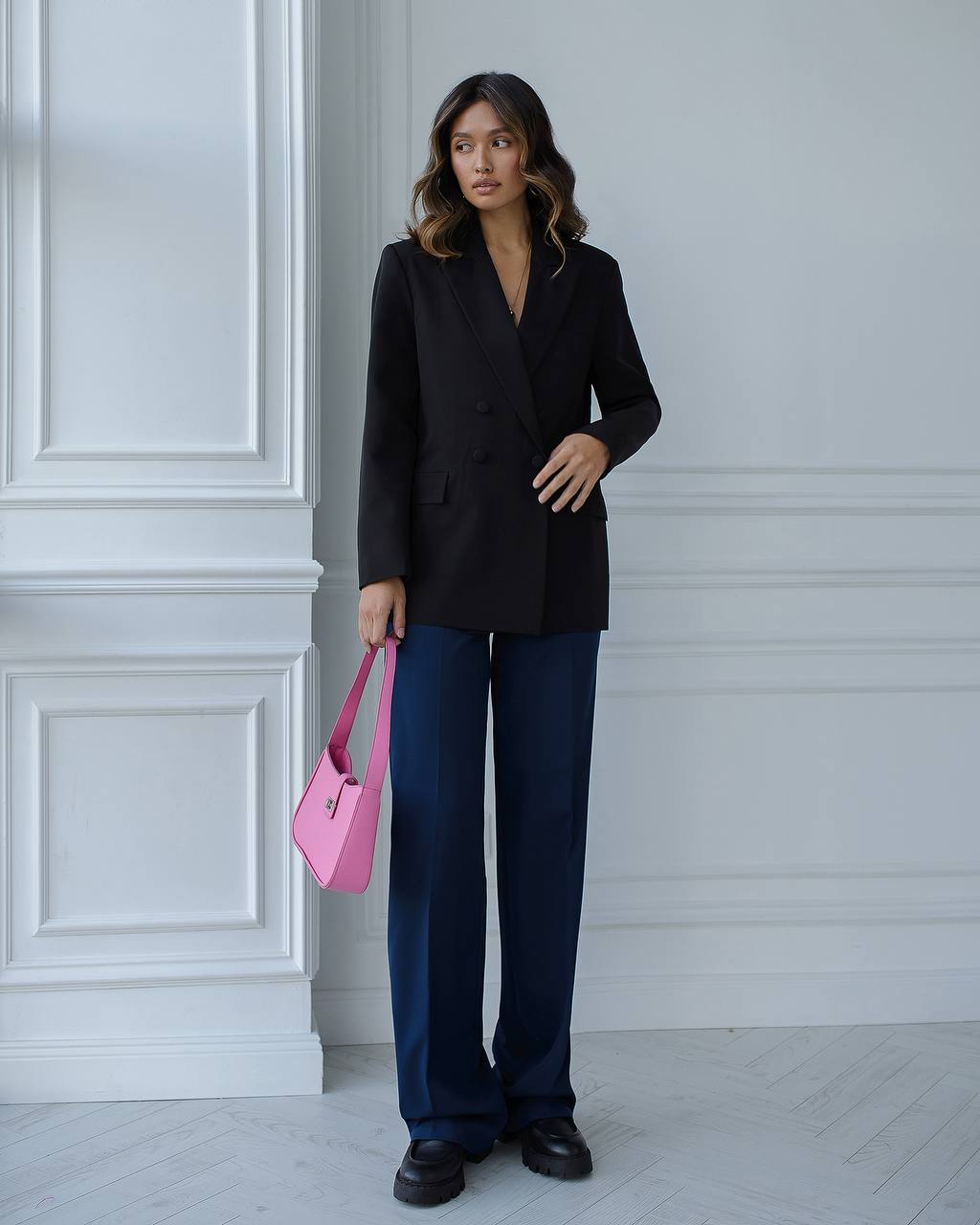Woman in black blazer and blue pants holding a pink bag against a white wall.