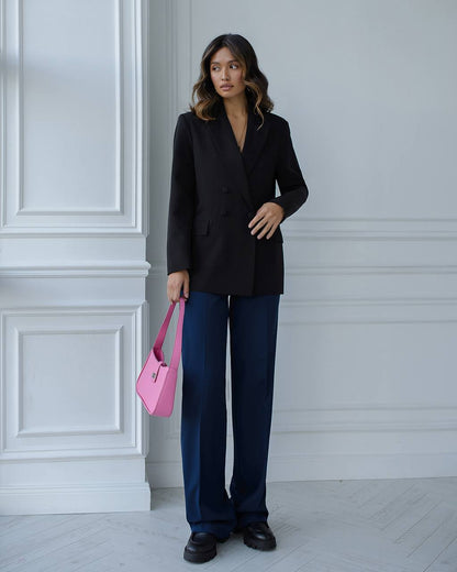 Woman in black blazer and blue pants holding a pink bag against a white wall.