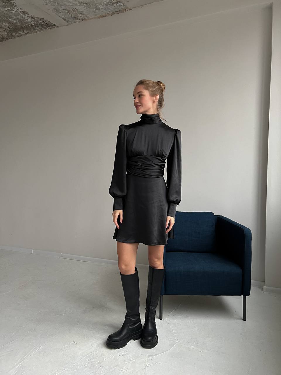 Woman in a black outfit standing in a minimalistic room with a blue chair.