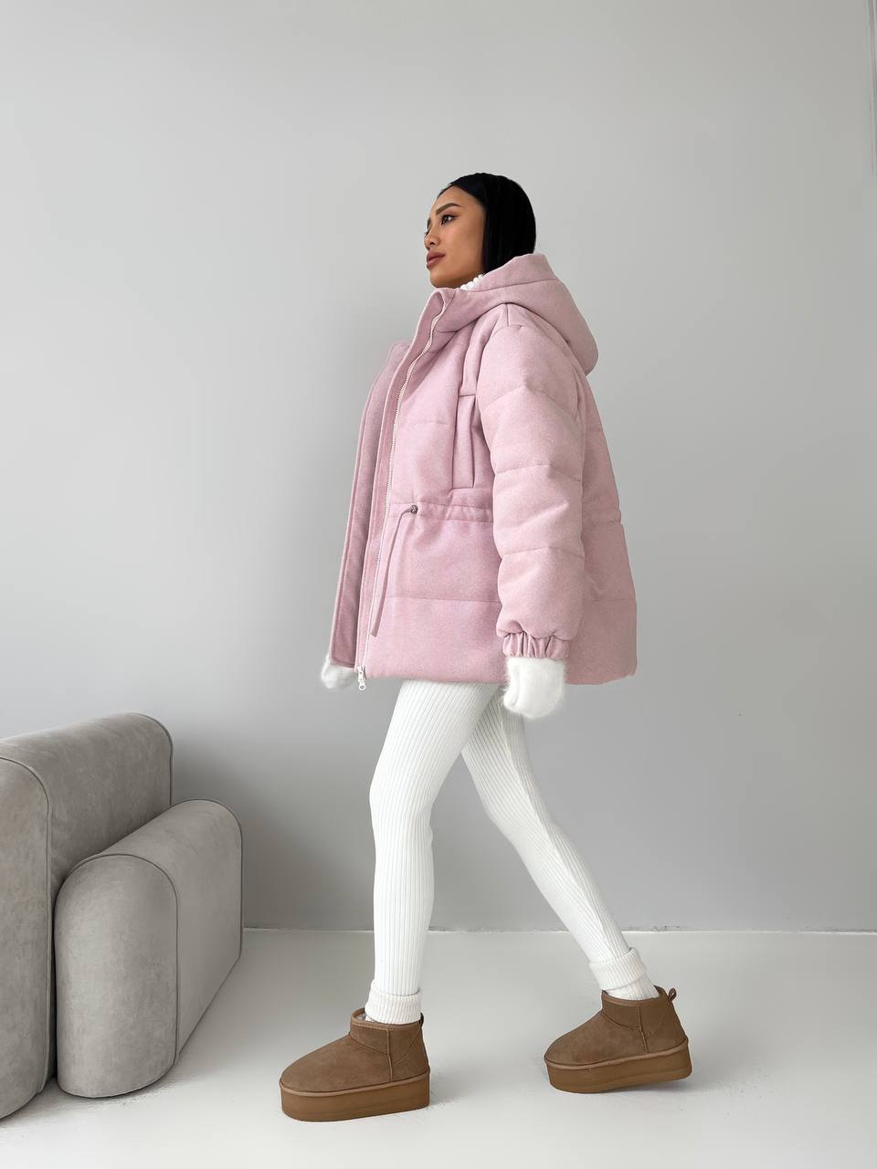 Person wearing a pink puffer jacket and white leggings standing in a minimalistic room.