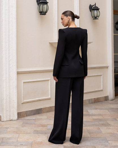 A woman stands outdoors in front of a cream-colored building wall, shown from the back wearing a tailored black long-sleeve blazer with structured shoulders and matching wide-leg trousers.