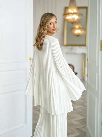 A woman wearing a white pleated long-sleeve top and matching pleated trousers, photographed indoors in a bright white room with gold-toned door handles and hanging lights in the background.
