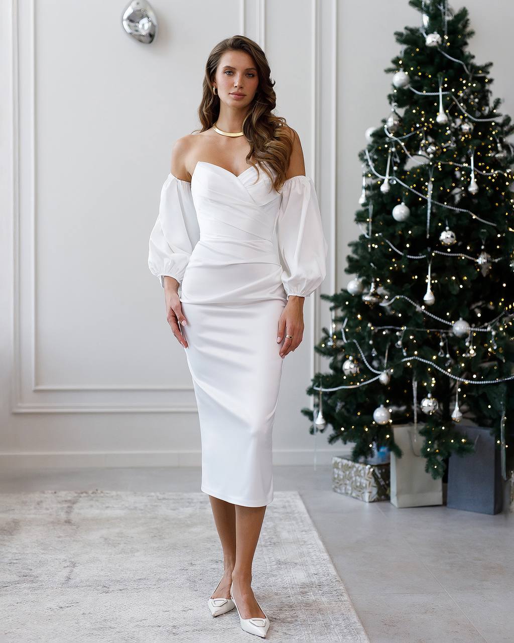 Woman in a white off-shoulder dress standing in a room with a decorated Christmas tree.