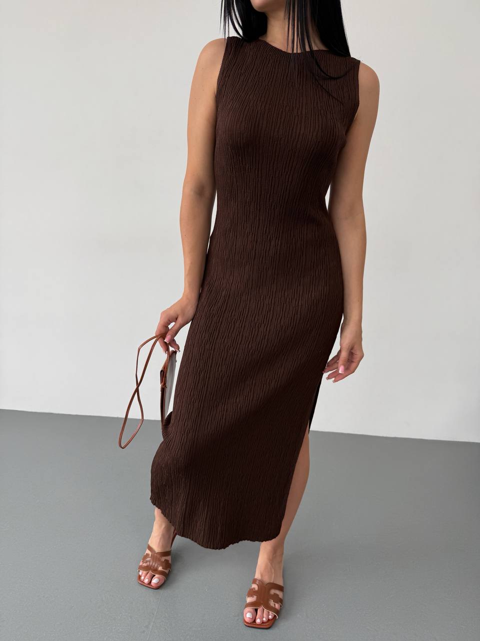 Woman wearing a brown sleeveless dress holding a matching handbag against a plain background