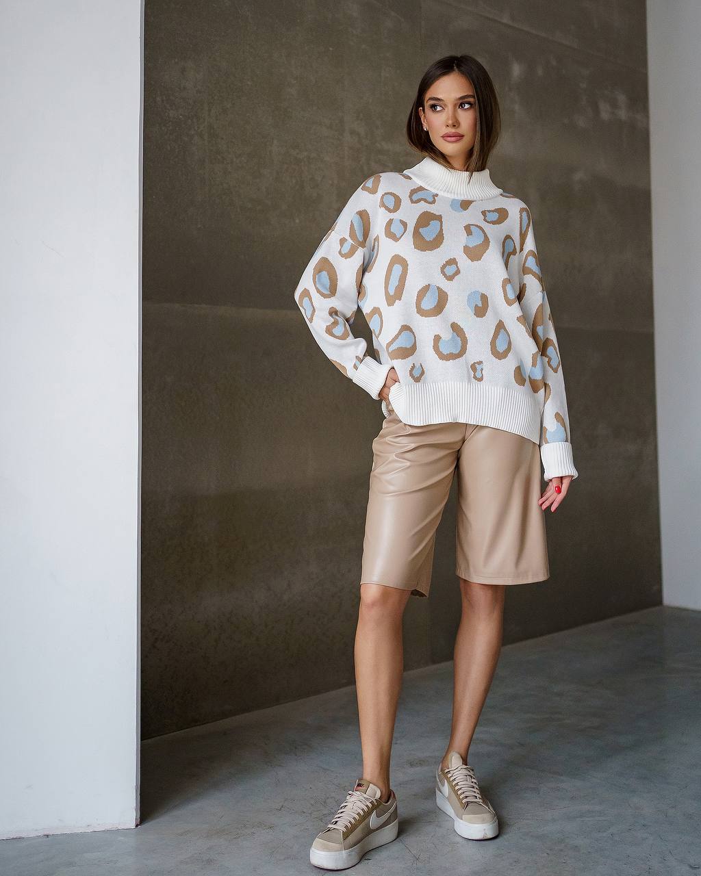 Woman wearing a patterned sweater and beige shorts against a neutral wall.