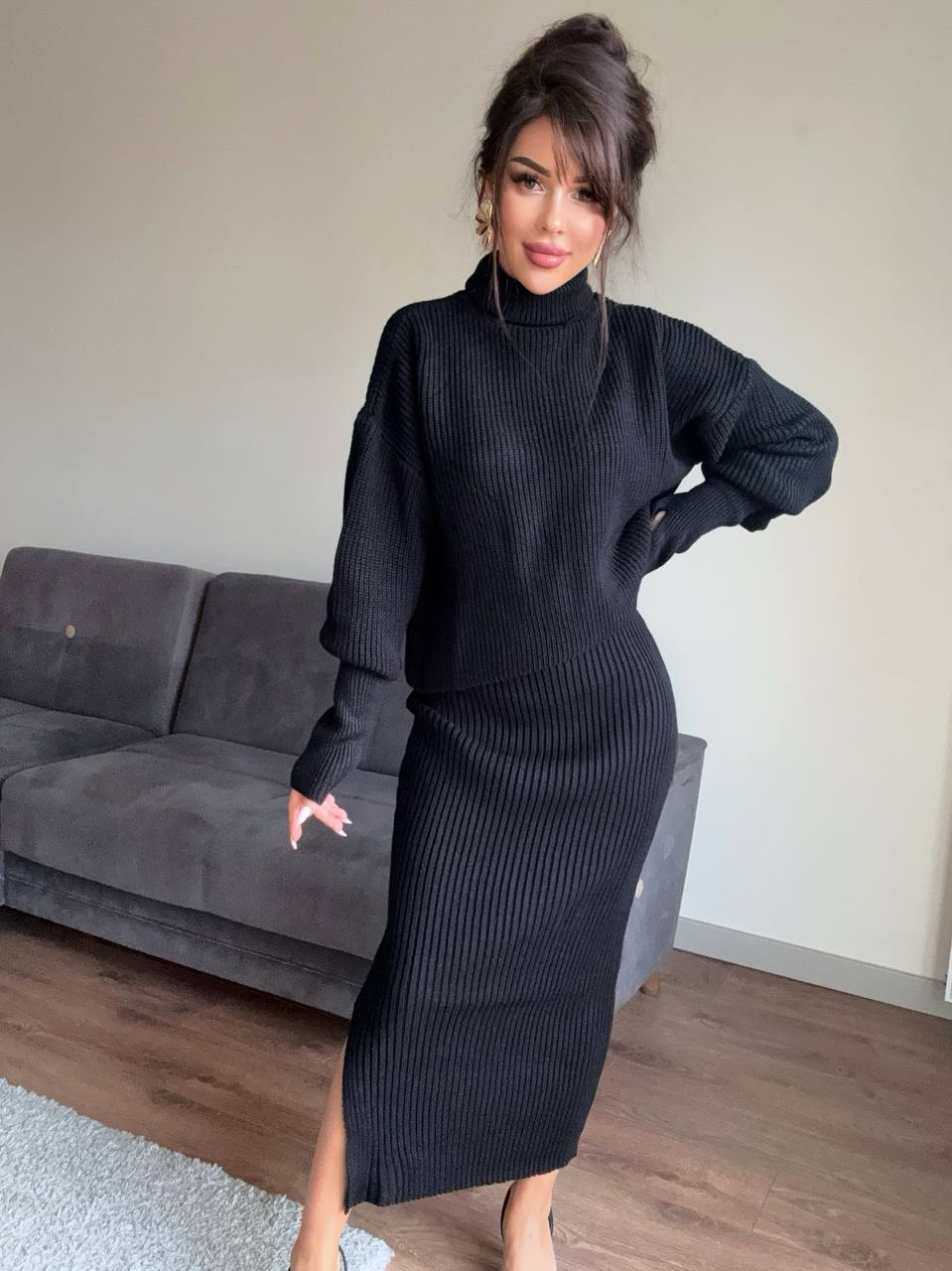 Woman wearing a black ribbed knit dress standing in a room with a gray couch.