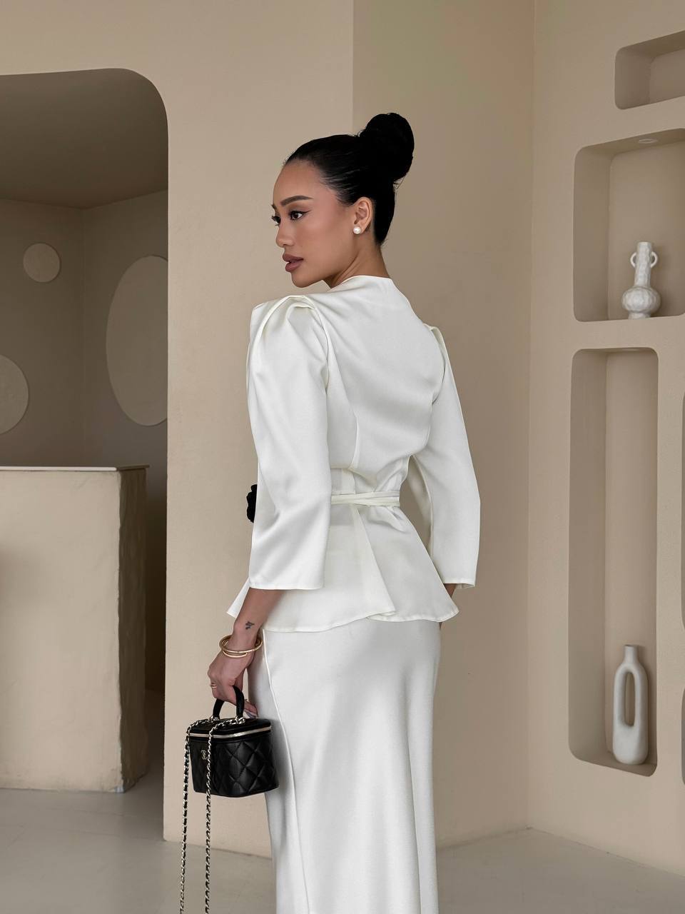 Woman in a white outfit holding a black handbag in a minimalistic setting.