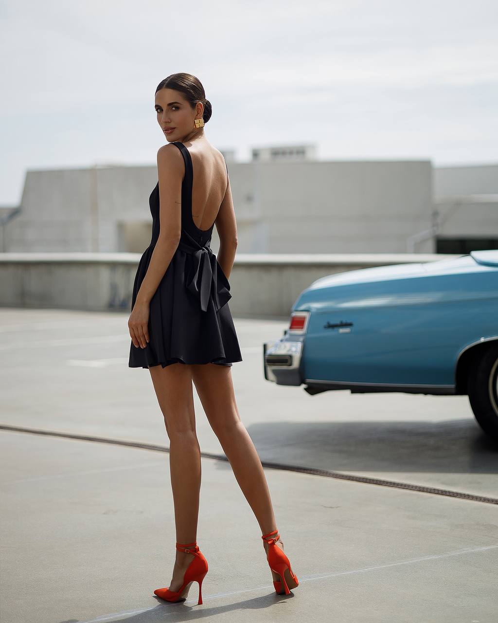 Woman in a black dress with an open back and red high heels standing in a parking lot with a blue car.