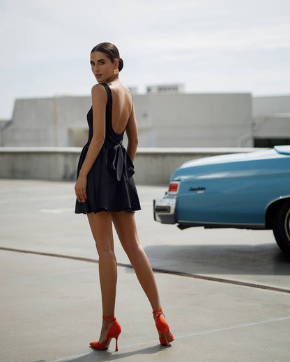 Woman in a black dress with an open back and red high heels standing in a parking lot with a blue car.