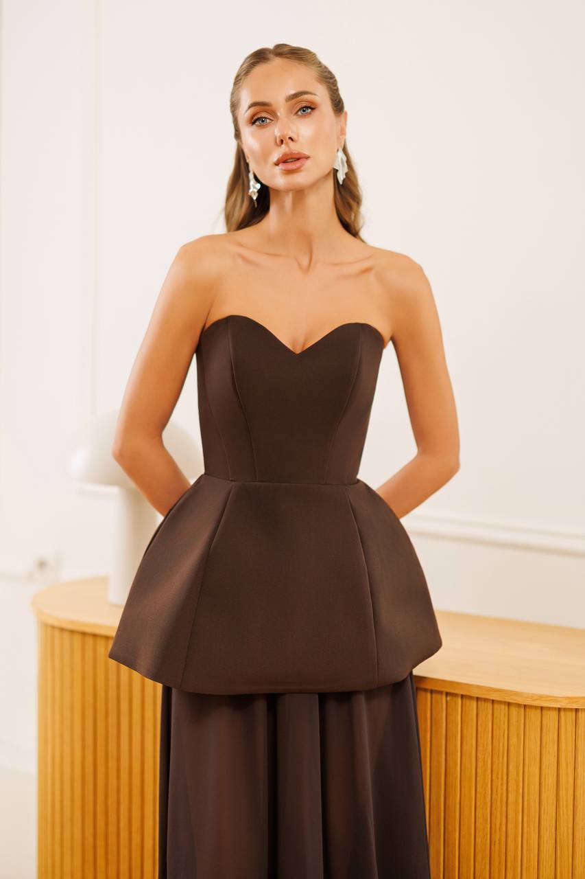 A woman modeling a strapless dark brown peplum dress with a sweetheart neckline and a flared waist, standing in front of a light-colored wall and wooden furniture.