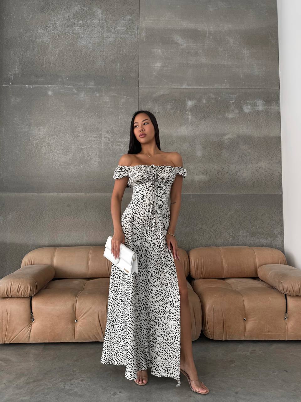 Woman in a patterned off-shoulder dress standing in front of a beige sofa against a concrete wall.