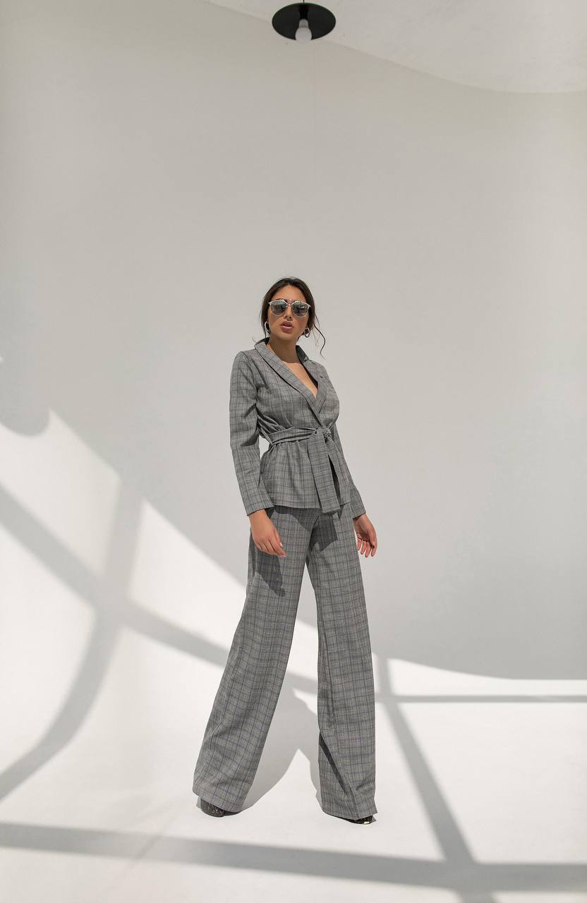 Woman wearing a gray checkered suit standing against a white wall with shadows.