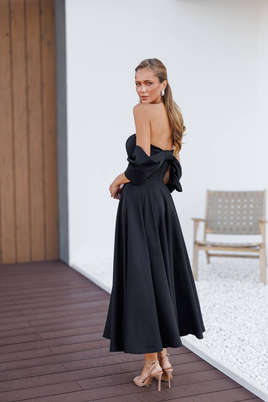 Woman in a black evening gown standing on a wooden deck with a white wall and chair in the background.