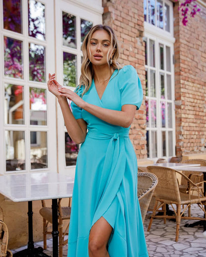 Woman in a turquoise dress standing in an outdoor cafe setting.