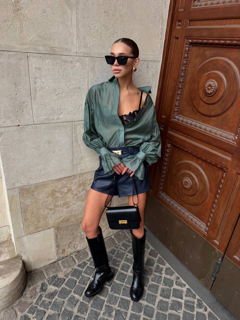Woman in green satin shirt and black boots standing next to a wooden door.