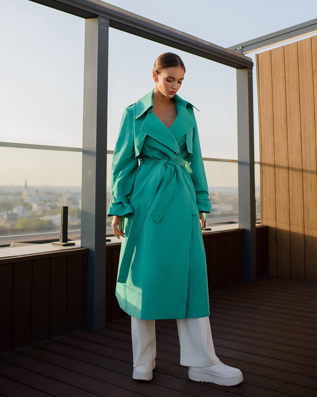 Woman in a teal trench coat standing on a rooftop with a cityscape view.