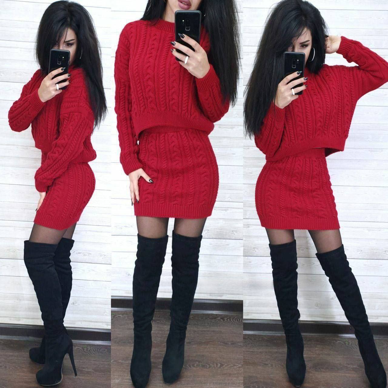 Woman wearing a red cable knit dress and black knee-high boots against a white brick wall.
