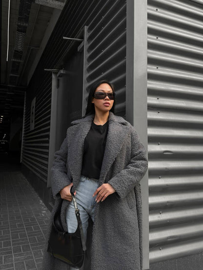 Person wearing a long gray coat and sunglasses standing against a corrugated metal wall.