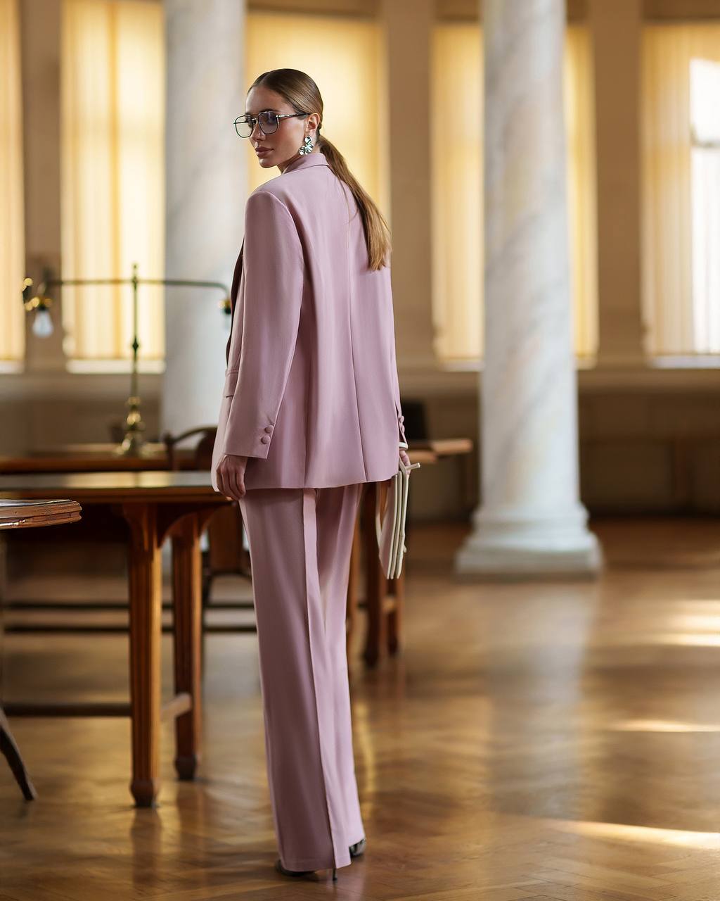 Woman in a pink suit standing in an elegant room with columns and wooden floors.