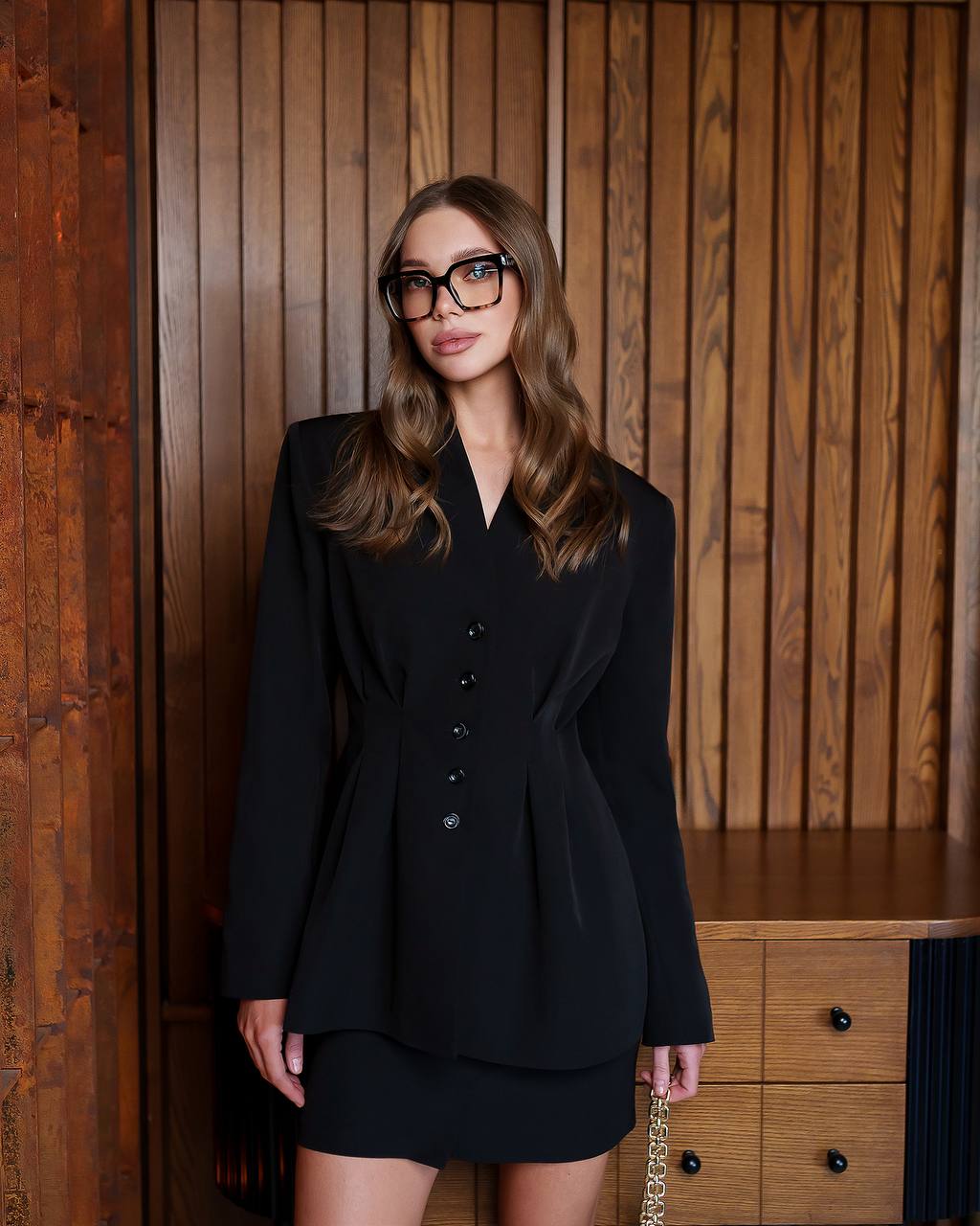Woman wearing a black outfit with glasses in front of a wooden panel background