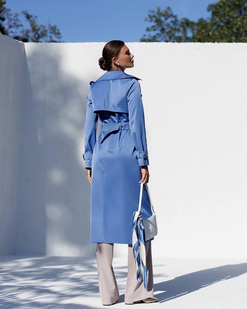 Woman wearing a blue trench coat standing against a white wall with a clear blue sky.