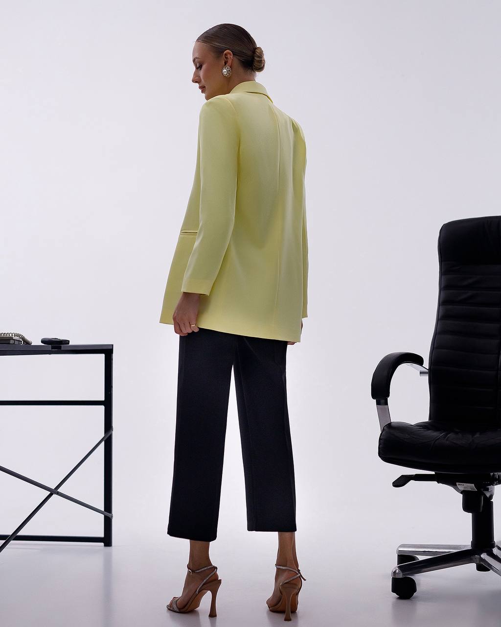 A woman stands in profile wearing a pale yellow blazer, black cropped trousers, and brown high-heeled sandals in a bright studio setting with a black office chair and small table visible.