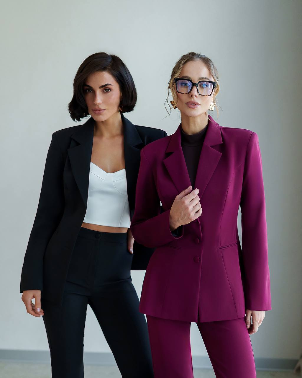 Two women wearing suits standing against a plain background