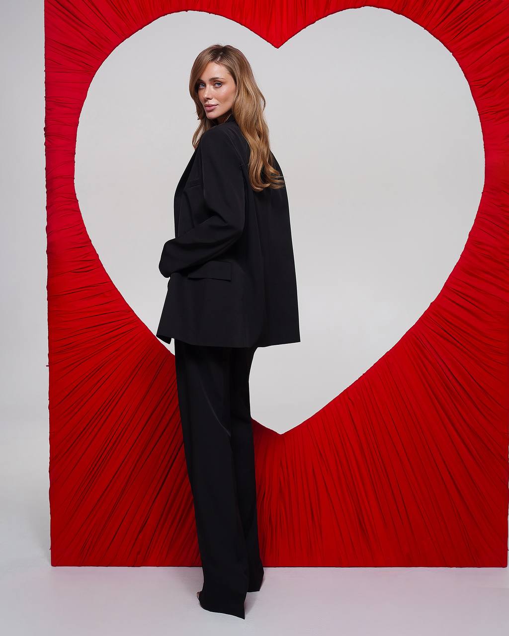 Person wearing a black suit standing in front of a red heart-shaped backdrop