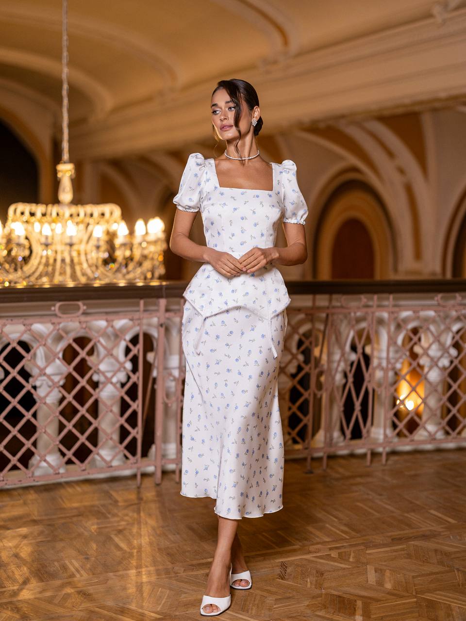 Woman in a white dress standing in an elegant indoor setting with chandelier and wooden floor.