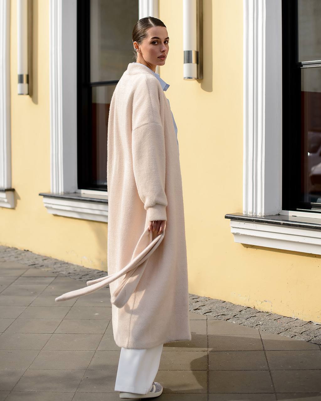 Woman wearing a long beige coat standing in front of a yellow building with large windows.