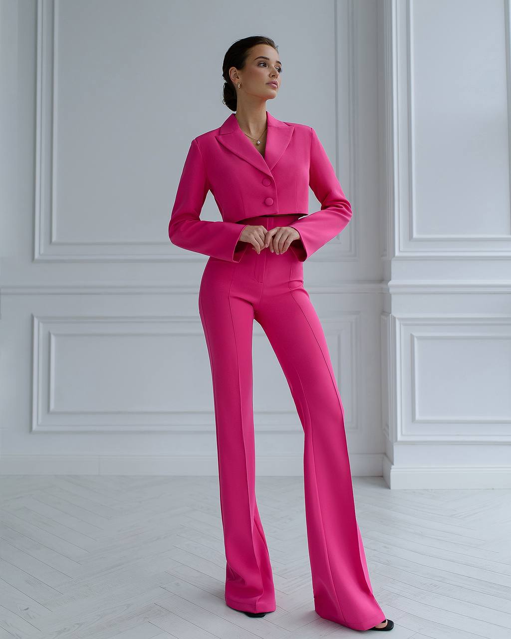 Woman wearing a bright pink suit standing against a white paneled wall.