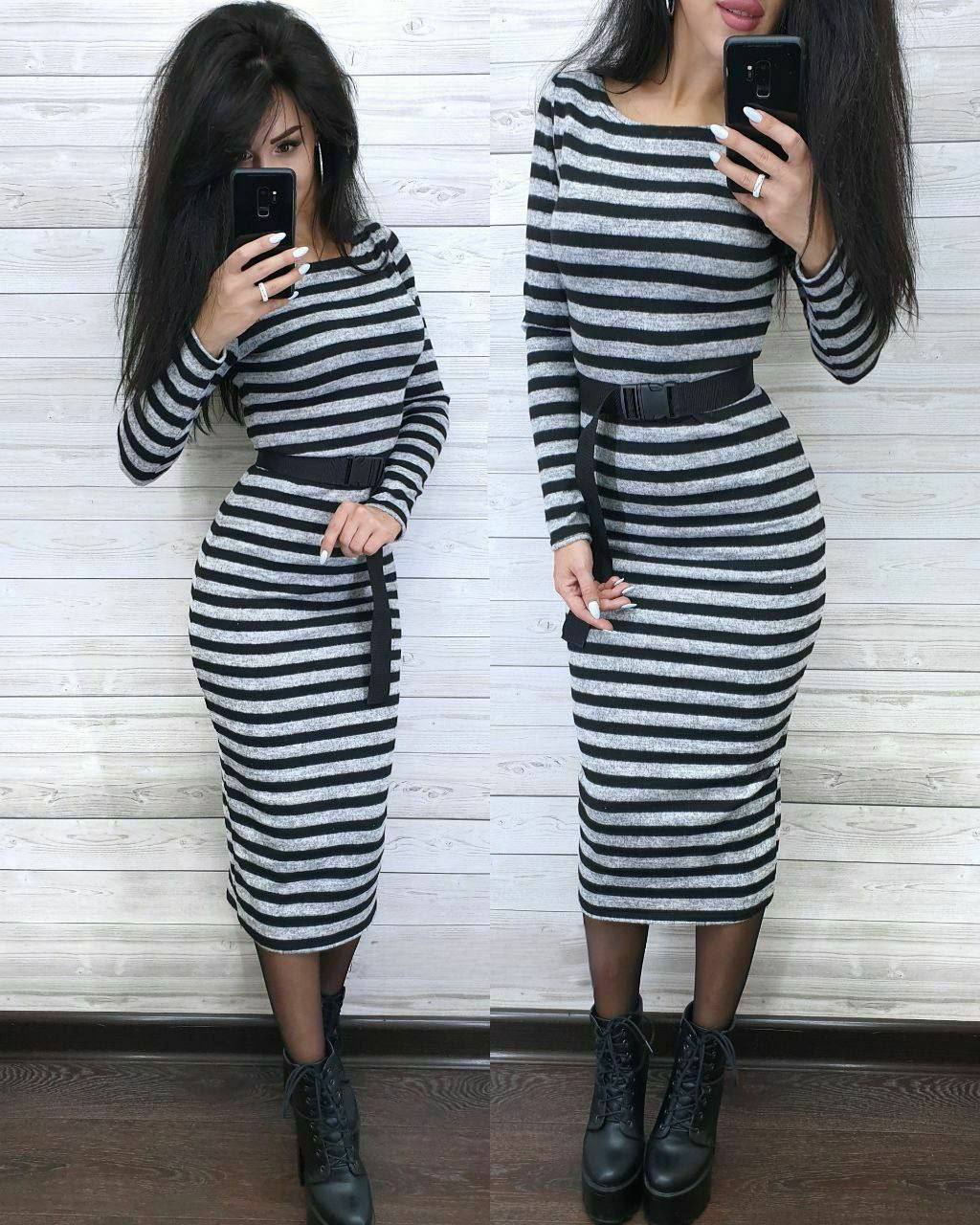 Person wearing a black and white striped dress with a belt, standing against a white wooden panel wall.