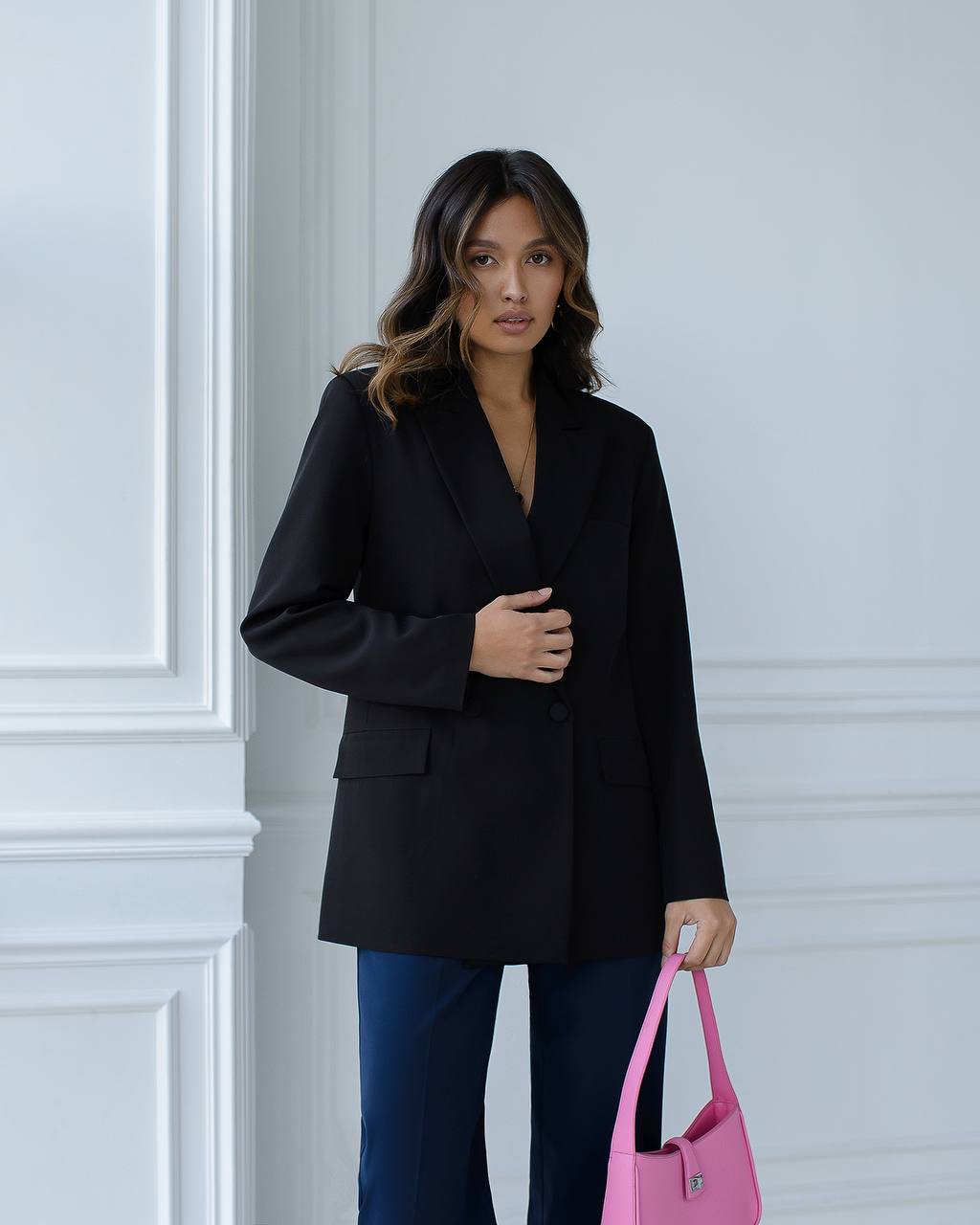 Woman wearing a black blazer and blue jeans, holding a pink handbag against a light-colored wall.