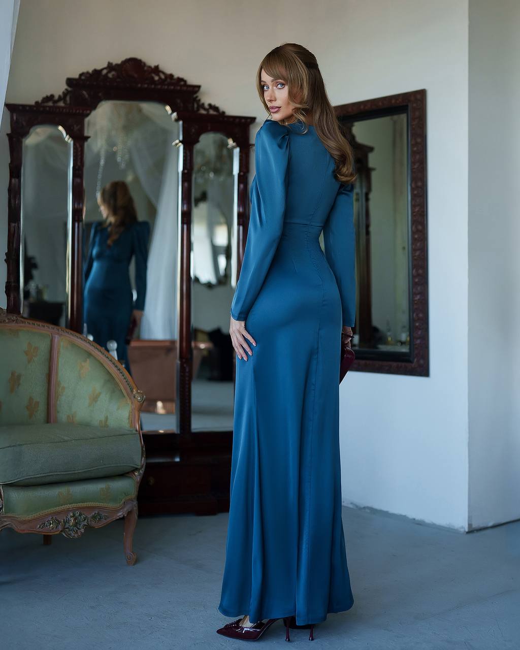 Woman in a blue dress standing in a room with vintage furniture and mirrors.